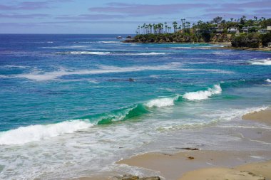 Dalgalar açık mavi gökyüzünün altında kumlu kumsala çarpıyor