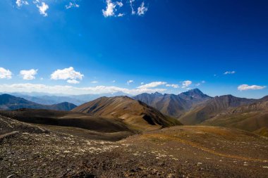 Geniş panoramik manzara parlak mavi gökyüzünün altındaki kayalık dağları gösteriyor.