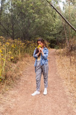 Siyah saçlı, kot ceketli genç beyaz bir kadın. Ormanın ortasında bir patikada sarı çiçekleri koklarken kameraya bakıyor. Etrafında bir sürü yeşillik var.