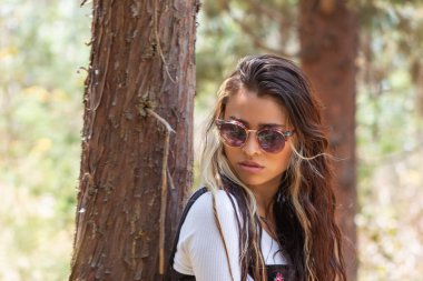 Güneş gözlüğü takan genç beyaz bir kadının bir ağaca yaslanıp güneşli bir günde ormanda yere ciddi ciddi baktığı bir fotoğraf..