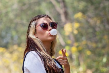 Güneş gözlüklü genç bir kadının, güneşli bir günde kameraya bakıp, sakız yapıp, kamerayı işaret edişinin orta boy kısa görüntüsü.