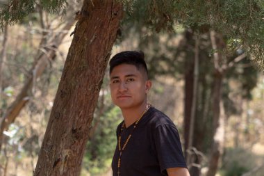 Güneşli bir günde, ormanda küçük bir gülümsemeyle kameraya bakan genç bir Latin 'in orta boy kısa çekimi.