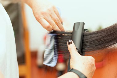 Tarağı ve saç düzleştiricisi kullanarak bir kadının uzun siyah saçını tarayan bir stilist.