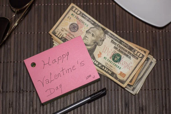 A small pink Valentine\'s note with a small wad of cash on a bamboo placemat surrounded by a pen, plate and sunglasses. Valentine\'s Day gift. Text: Happy Valentine\'s Day, Love You.