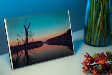A landscape photograph next to a vase and some flowers on a table inside a room at night. Hingham, Massachusetts Picture.