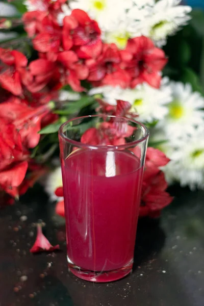 Red juice in a glass with defocused red and white flowers in the background