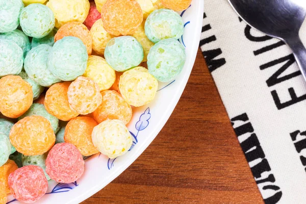 Close up overhead shot of a bowl of colorful sugary cereal next to a cloth napkin and spoon on a wooden table