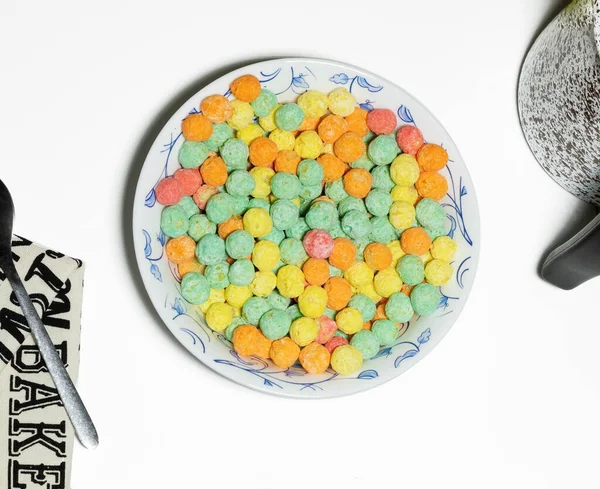 Overhead shot of a bowl of colorful sugary cereal next to a cloth napkin, spoon and small pot on a white background