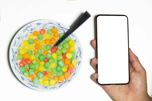 Overhead shot of a man holding a cell phone with a blank screen next to a bowl of colorful sugary cereal with milk on a white background