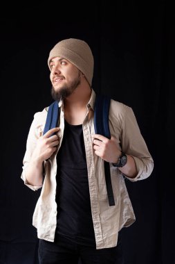 Bearded young college man smiling on a black background while holding his backpack