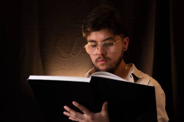 A young man with glasses reading a book in front of a dark background