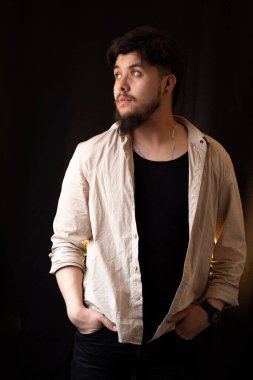 A young white man with light eyes and a beard standing in front of a black background