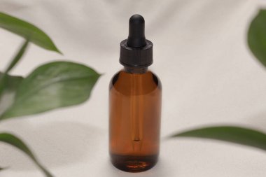 Arka planda yeşil yapraklı, bej renkli bir damlacık şişesi.