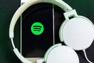 Bogota, Kolombiya - 26 Temmuz 2023. Siyah arka plandaki Spotify uygulamasını kullanarak beyaz kulaklıklara bağlı bir akıllı telefon. Yeşil ışık. Eğlence uygulamaları.