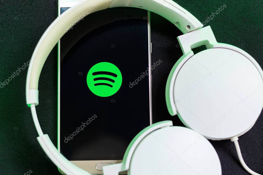 Bogota, Colombia - July 26, 2023. A smartphone connected to white headphones using the Spotify app on a black background. Green light. Entertainment apps.