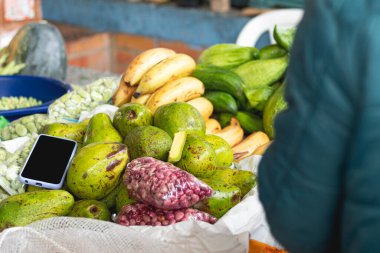 Yerel bir çiftçi dükkanındaki avokadoların üzerinde duran bir cep telefonu. Muz, fasulye ve taze organik ürünler. Çiftçi pazarı
