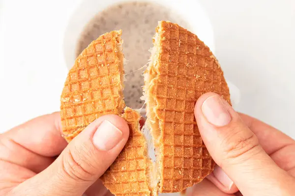 Point of view of a person breaking a stroopwafel in half with a cup of coffee out of focus in the background. Tea time. Caramel filling.Traditional Dutch food concept.
