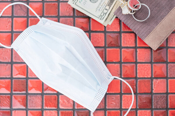 Close-up overhead view of a face mask next to a wallet, bills and keys on a red grid tile background. Elements to go out of the house.