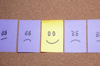 Kızgın yüzleri mantar panoya dizilmiş post-it notları, ortasında mutlu yüz kontrastı olan bir not. Ofiste duygular.