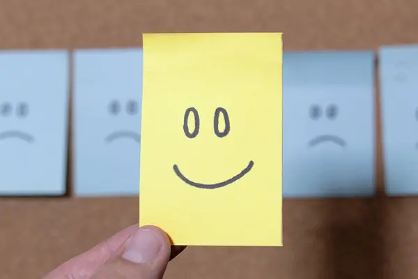 Bir insan mavi notaların önünde sarı yapışkan bir nota tutar ve üzgün yüzler bir mantar panoya yapışır..