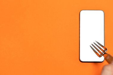 Boş ekranlı bir cep telefonunun çatalını tutan bir el. Turuncu arka plan. İnternetten yemek alışverişi. Kopyalama alanı olan resim.