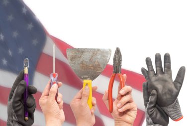 İşçilerin elleri, güvenlik eldiveni takanların bazıları iş aletlerini tutuyor. Arka planda ABD bayrağı bulanık. İşçi Bayramı konsepti.