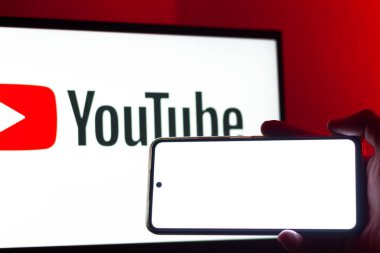 Bogota, Kolombiya - 12 Temmuz 2024: Bir el, youtube logosunun bulunduğu büyük ekranın önünde boş ekranlı bir akıllı telefonu tutuyor.
