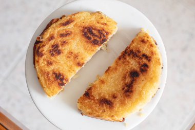 Izgara peynirli arepanın üst görüntüsü, altın kahverengi kenarları ve çıtır çıtır dokuları beyaz bir tabakta servis ediliyor, ikiye bölünmüş ve yemeye hazır. Geleneksel Kolombiya gıda kavramı