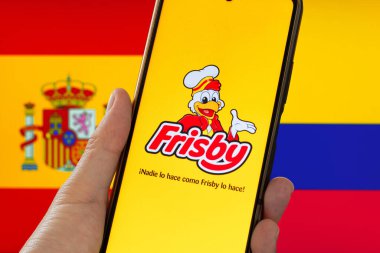 Bogota, Kolombiya - 14 Mayıs 2025. İspanya ve Kolombiya 'nın bayrakları önünde Frisby uygulamasının açık olduğu bir cep telefonu, markayla ilgili uluslararası anlaşmazlığı ima ediyor. Kolombiya kökenli restoran zinciri.