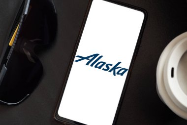 Bogota, Kolombiya - 13 Mayıs 2025: Koyu gözlükler ve kapaklı bir kahve fincanı Amerika Birleşik Devletleri 'nden bir havayolu şirketi olan Alaska Havayolları' nın logosunu gösteren bir cep telefonu