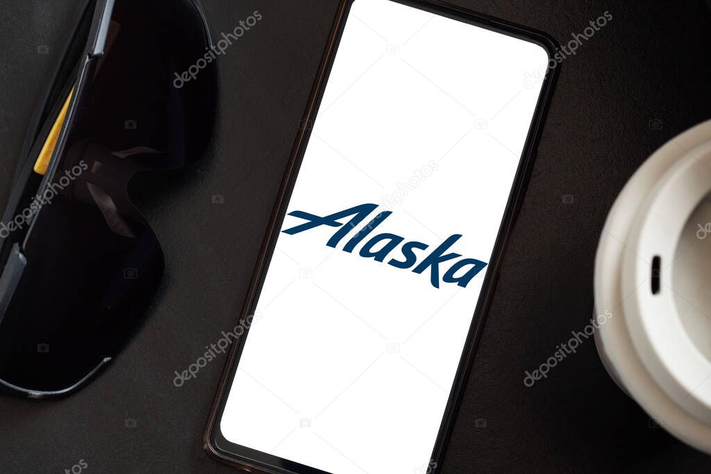 Bogota, Colombia - May 13, 2025: Overhead view of dark glasses and a coffee cup with lid accompanying a cell phone showing the logo of Alaska Airlines, an airline from the United States