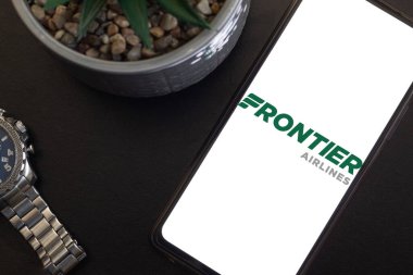 Bogota, Kolombiya - 13 Mayıs 2025: Frontier Airlines logosu, ABD düşük maliyetli havayolu, cep telefonu ekranında karanlık masaüstünde sulu bitki ve metal saat, seyahat ve kişisel planlama kavramı
