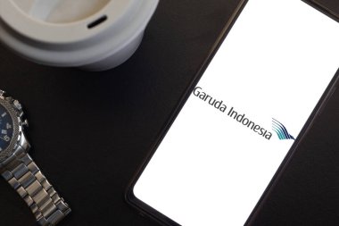 Bogota, Kolombiya - 13 Mayıs 2025: Endonezya 'nın ulusal havayolu şirketi Garuda Endonezya, cep telefonunda, koyu bir arka plan, uluslararası uçuş teması ve yürütme tarzında kahve ve saatin yanında