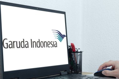 Bogota, Kolombiya - 14 Mayıs 2025: Garuda Endonezya logosu, Endonezya 'nın ulusal havayolu, bazı kalemler ve ellerle birlikte bir fare, gayri resmi ofis ortamı ve çevrimiçi uçuş rezervasyonu kullanarak bilgisayarda görünür.