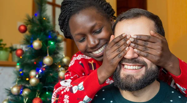 Happy african woman covering eyes of bearded man with her hands happy receiving a christmas gift in front of the christmas tree wearing christmas sweaters