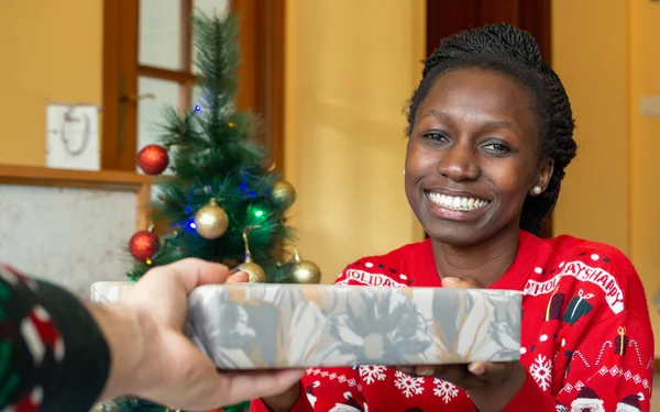 Young african woman receiving a wrapped gift in front of a Christmas tree wearing a christmas sweater Looking at camera