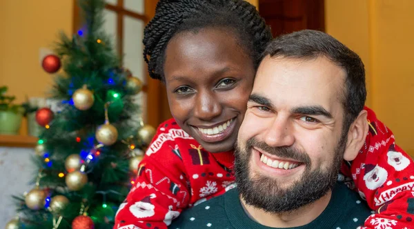 Farklı ırktan çiftler Noel 'in tadını çıkarıyorlar. Süslü bir ağacın önünde Noel süveteri giyerken kameraya bakıyorlar..