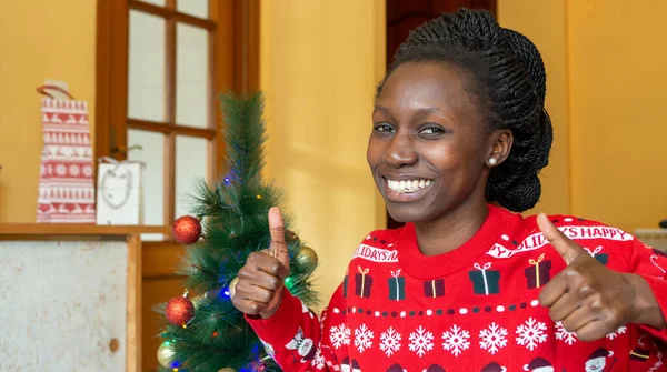 Happy young african in front of the christmas tree wearing a christmas sweater Looking at camera. Thumbs up