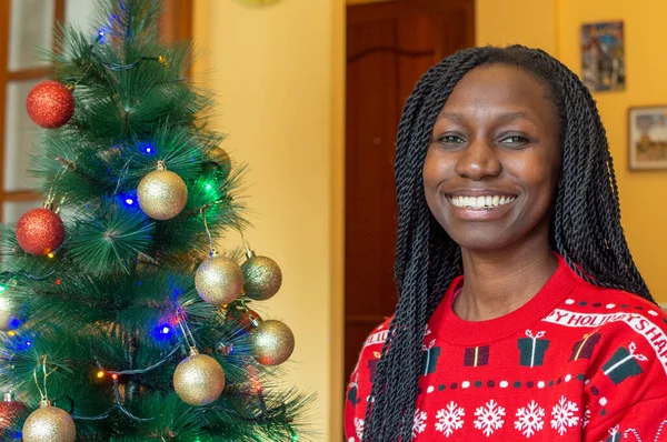 Happy young african in front of the christmas tree wearing a christmas sweater Looking at camera