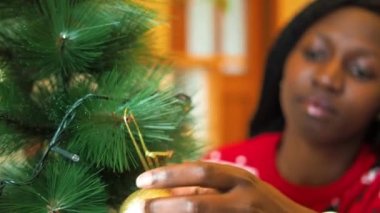 Mutlu, genç Afrikalı bir kadın Noel ağacına altın bir top koyuyor Noel süveteri giyiyor.