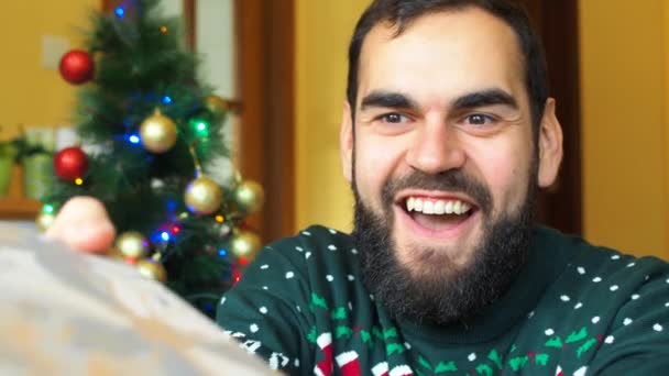 一个满脸胡须的快乐男人兴奋地在圣诞树前收到包装好的礼物 身穿圣诞毛衣 — 图库视频影像