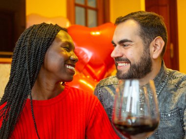 portrait of an interracial couple looking at each other laughing with glasses of red wine enjoying valentines day