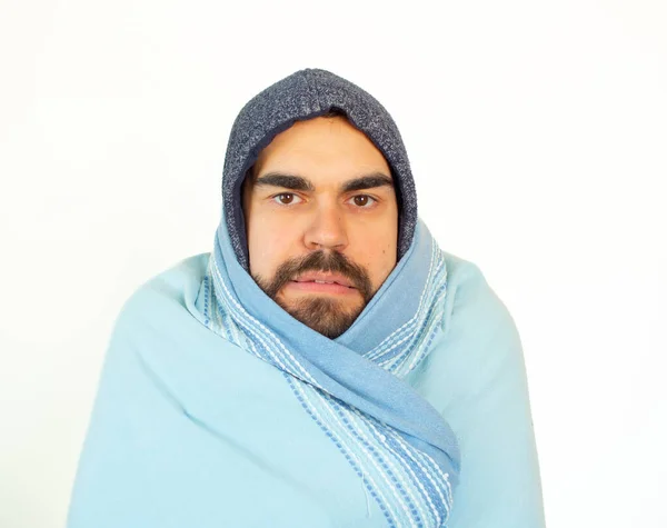 Young bearded man wearing winter clothes covering with a blue blanket. Cold expression. Looking at camera. Isolated white background.