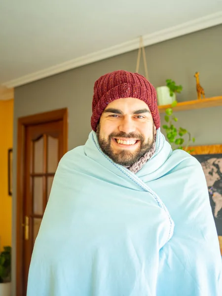 Young bearded man covering himself with a blanket to fight cold wave. Wearing winter clothes in a cozy living room. Looking at camera.