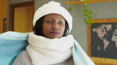 Young woman covering herself with a blanket to fight cold wave. Wearing winter clothes in a cozy living room. Looking at camera.