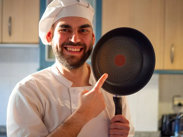 portrait of a young bearded chef smiling and pointing a non stick pan looking at camera