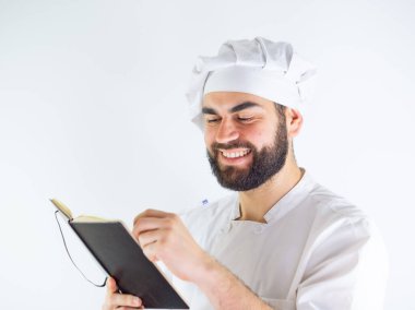 Genç aşçı bir defter kullanıyor, yemek tarifi yazıyor. Beyaz bir arkaplanda izole edilmiş