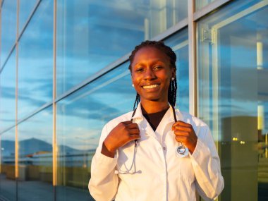 Genç bayan doktor kameraya bakarak hastanenin önünde boynuna steteskop takıyor.