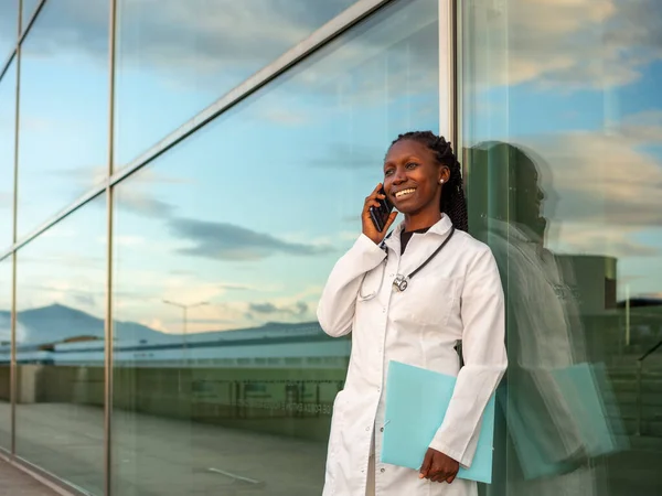 young female doctor talking by phone outdoors in front of a hospital