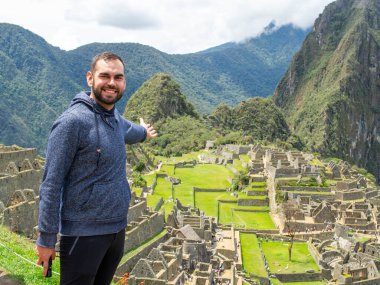 A Happy bearded traveler pointing to Machu Picchu wonders in a sunny day clipart